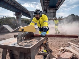 Arbeiter in gelber Schutzkleidung bedient Tischsaege auf einer staubigen Baustelle unter einer Bruecke