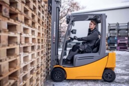 Ein Arbeiter in schwarzer Arbeitskleidung faehrt Gabelstapler neben einem hohen Stapel Holzpaletten im Freien