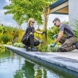 Zwei Personen, ausgestattet mit Sicherheitskleidung und -brillen von uvex, arbeiten in einem grünen Garten