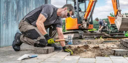 Ein Arbeiter, der Sicherheitskleidung, -handschuhe und -brille von uvex trägt, verlegt Pflastersteine mit einem Hammer auf einer Baustelle