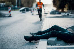 Eine Person in orangefarbener Warnweste im Hintergrund, während im Vordergrund die Beine einer auf dem Boden liegenden Person zu sehen sind.